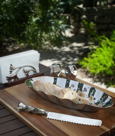 ginkgo napkin holder is displayed on table with napkins and tray, salt and pepper, and bread knife among it. trees and bushes with a trail path is shown in the background.